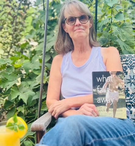 Picture of Karin Anderson sitting outside in a green lanscape holding her book, What Falls Away 