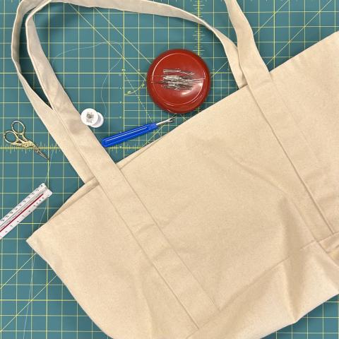 A natural-color canvas tote bag lays on a green gridded cutting mat, surrounded by sewing notions (thread, seam ripper, ruler, and pins)